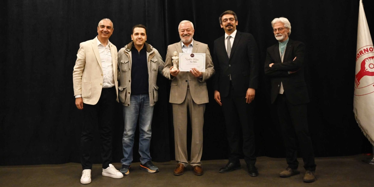 Siyah bir perde önünde yan yana duran beş erkek, bir ödül töreninde poz veriyor. Ortada duran kişi, elinde küçük bir büst ve üzerinde 