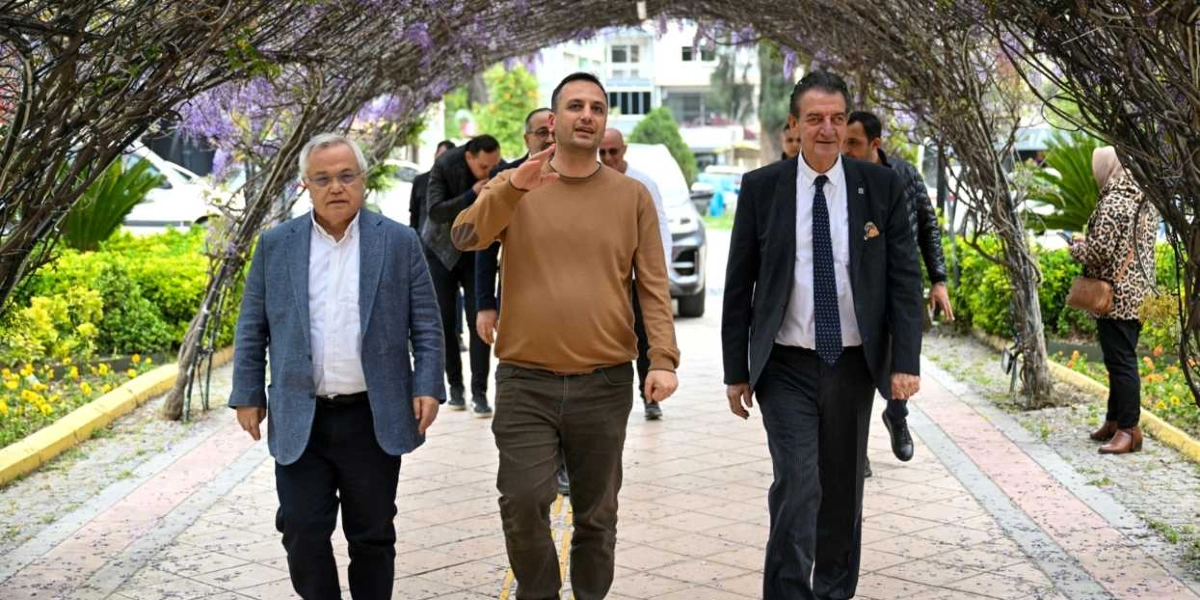 Mor çiçekli sarmaşıklarla kaplı bir tünelin altında yürüyen üç erkek; solda gri ceketli, ortada kahverengi kazaklı ve sağda lacivert takım elbiseli kişiler, yanlarında diğer katılımcılarla birlikte bir park yolunda ilerliyor.