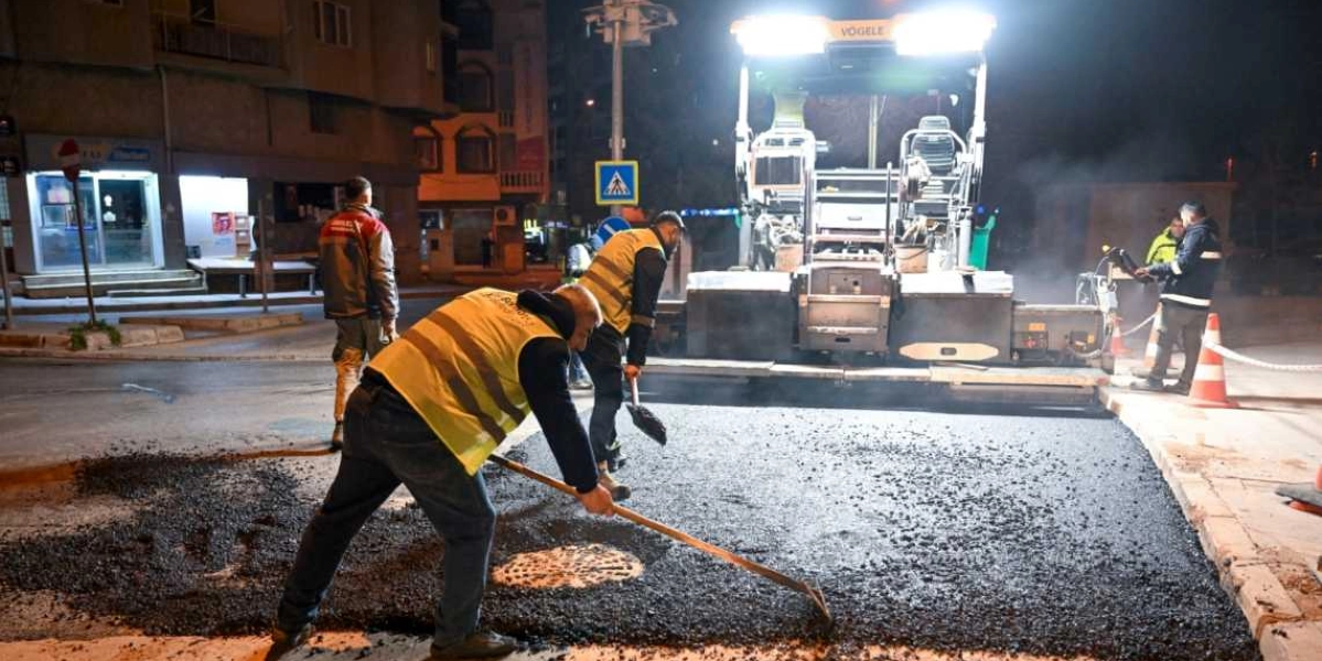Gece vakti Bornova'da bir sokakta çalışan belediye işçileri, önlerinde duran büyük bir asfalt serme makinesiyle yol yenileme çalışması yapıyor. Sarı reflektörlü yelekler giyen işçilerden biri elindeki tırmıkla dökülen sıcak asfaltı düzeltirken, diğerleri makinenin çevresinde çalışmaya devam ediyor. Çalışma alanı turuncu trafik konileriyle çevrelenmiş durumda.