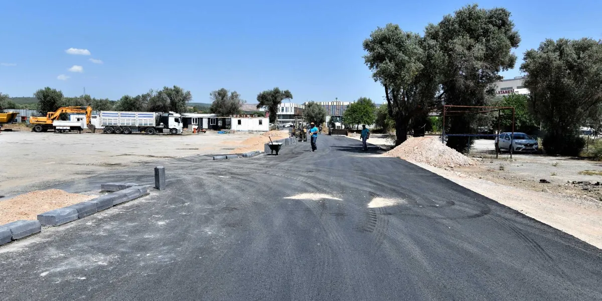 Açık havada, yeni asfaltlanmış geniş bir yol çalışması görülüyor. Yolun kenarlarında beton bordür taşları dizilmiş durumda. Arka planda bir kamyon, iş makineleri, konteynerler ve ağaçlar yer alıyor. Yol üzerinde yürüyen iki işçi ve sağ tarafta park halinde beyaz bir binek otomobil bulunuyor.