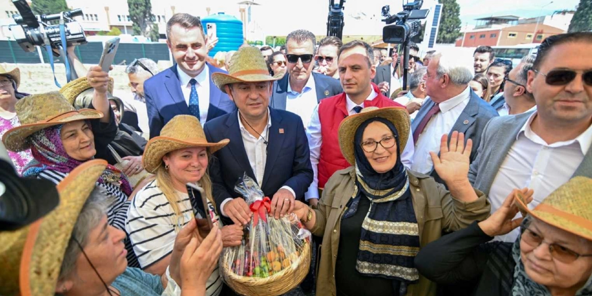 CHP Genel Başkanı Özgür Özel, Bornova'daki bir etkinlikte hasır şapka takan vatandaşlarla bir arada bulunuyor. Özel, elinde meyve dolu bir hediye sepeti tutarken, çevresindeki kadınlar ve diğer katılımcılarla birlikte gülümsüyor. Arka planda basın mensupları ve diğer yetkililer yer alıyor.