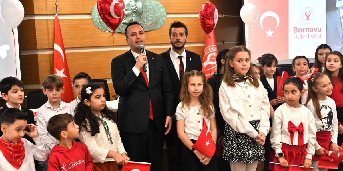 Bornova Belediye Başkanı, elinde mikrofonla konuşma yaparken yanında bir grup çocukla birlikte sahnede duruyor. Arka planda Bornova Belediyesi logosu, Türk bayrakları ve Atatürk silüeti yer alıyor. Çocukların birçoğu ellerinde küçük Türk bayrakları tutuyor, bazıları ise üzerinde Atatürk baskılı tişörtler giyiyor. Ortam, 23 Nisan Ulusal Egemenlik ve Çocuk Bayramı kutlaması atmosferini yansıtıyor.