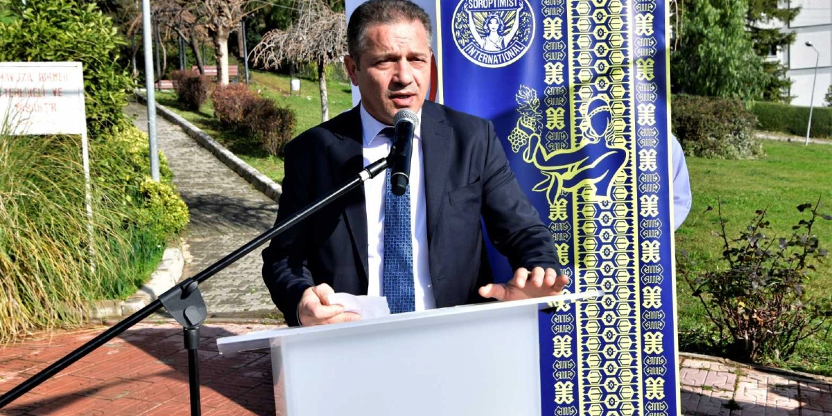 Koyu renk takım elbiseli ve mavi desenli kravatlı bir erkek, dış mekanda, üzerinde Soroptimist International logosu ve geleneksel motifler bulunan mavi bir pankartın önünde, kürsüde konuşma yapıyor.