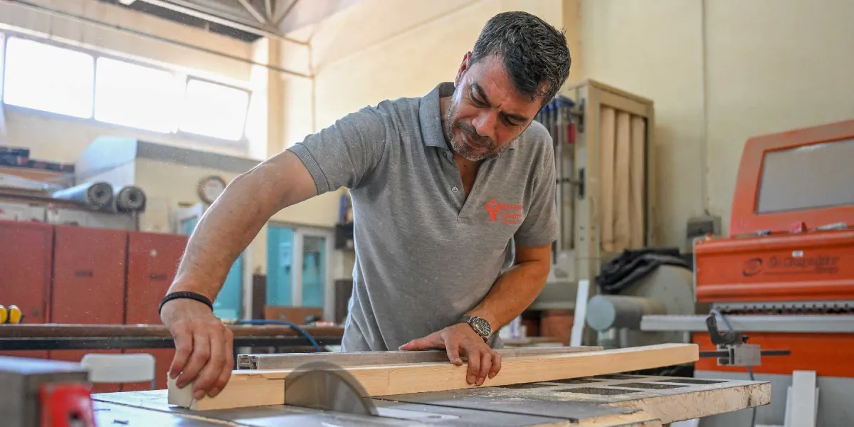 Bornova Belediyesi atölyesinde çalışan bir marangoz, tezgah üzerindeki ahşap bir kalası dairesel testere ile kesiyor.
