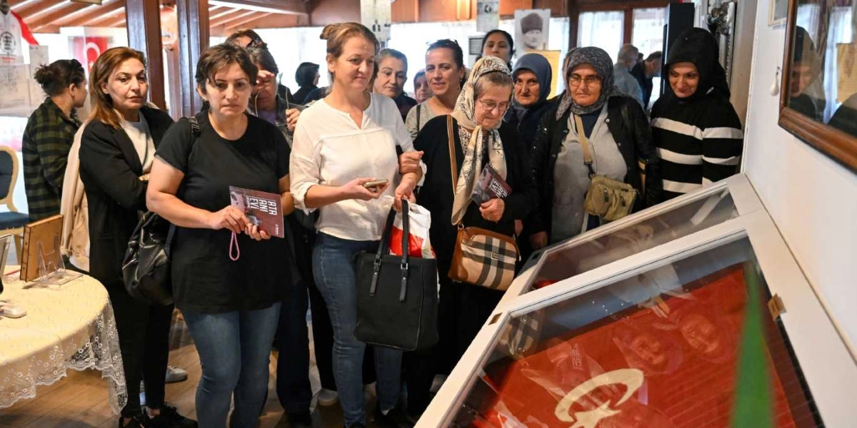 Belkahve'deki bir müze veya anı evinde, cam bir vitrin içerisinde sergilenen Türk bayrağını ilgiyle inceleyen bir grup kadın. Kadınlar, ellerinde 