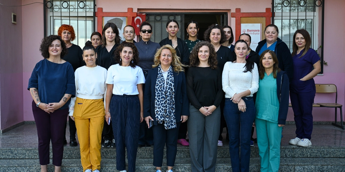 Bornova Belediyesi binasının girişinde, merdivenlerde yan yana duran, çoğunluğu sağlık çalışanı olduğu anlaşılan 17 kişilik kadın grubu. Arka planda pembe duvarlı bina, Türk bayrağı ve Bornova Belediyesi logosunun yer aldığı bir tabela bulunuyor. Kadınlar günlük kıyafetler ve bazıları sağlık personeli formalarıyla objektife gülümseyerek bakıyor.