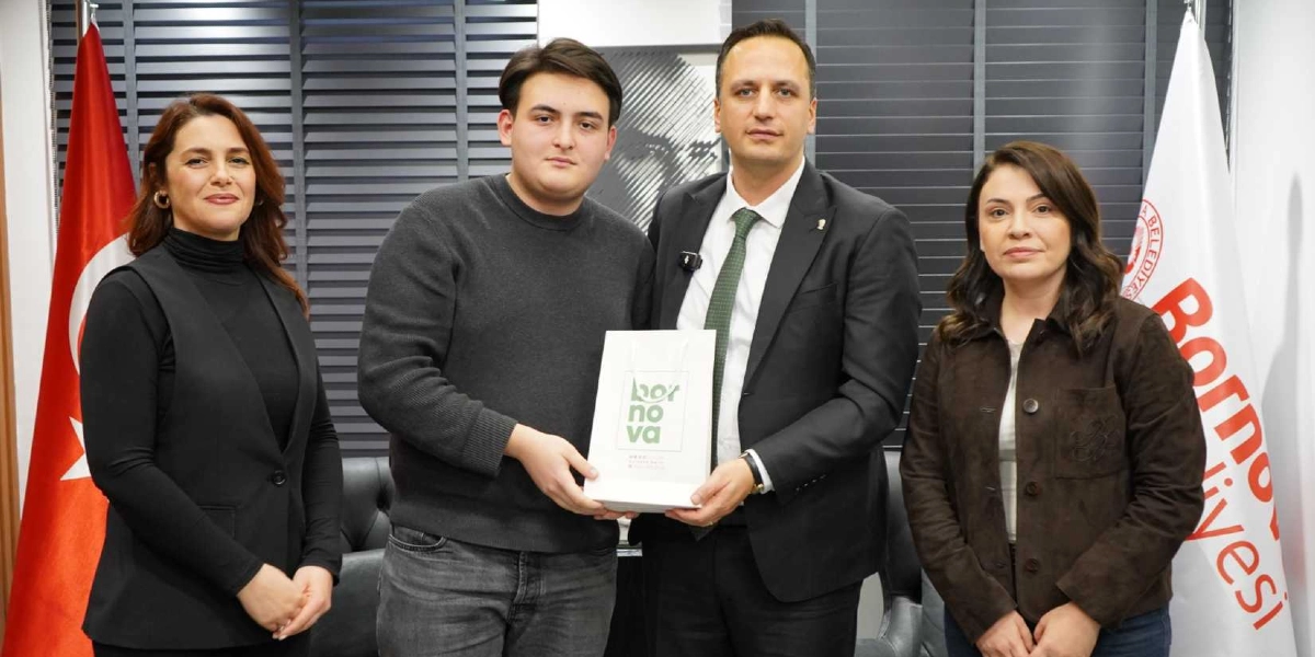 Bornova Belediye Başkanı, yanında iki kadınla birlikte başarılı bir öğrenciye üzerinde Bornova Belediyesi logosu bulunan bir hediye çantası takdim ediyor. Arka planda Türk bayrağı ve Bornova Belediyesi flaması yer alıyor.