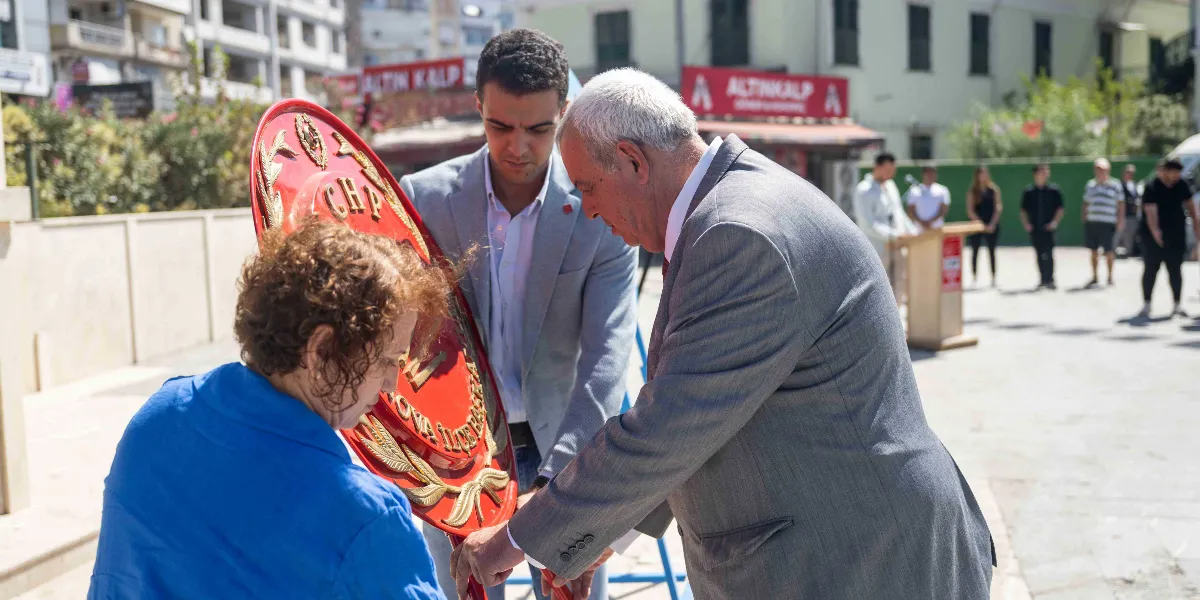 Açık havada düzenlenen bir törende, gri takım elbiseli bir erkek ve mavi kıyafetli bir kadın, üzerinde 