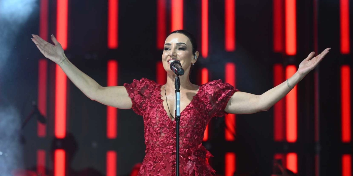 Kırmızı çiçek detaylı bir elbise giymiş kadın şarkıcı, sahnede mikrofon başında kollarını iki yana açarak şarkı söylüyor. Arka planda dikey kırmızı ışık şeritlerinden oluşan bir sahne dekoru bulunuyor.