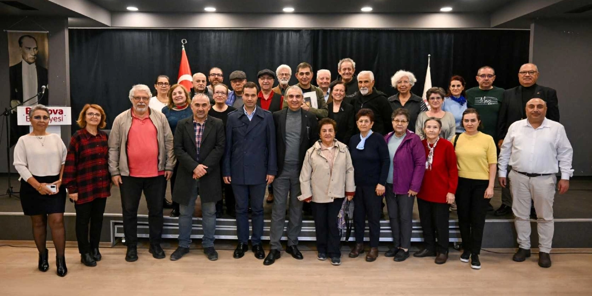 Kapalı bir salonda, sahnede yan yana duran yaklaşık otuz kişilik bir grup insan. Arka planda siyah bir perde, Türk bayrağı ve sol tarafta Atatürk portresi ile Bornova Belediyesi yazılı bir kürsü bulunuyor. Gündelik kıyafetler içindeki katılımcılar, bir söyleşi veya etkinlik sonrasında hatıra fotoğrafı çektirmek için objektife gülümseyerek bakıyor.