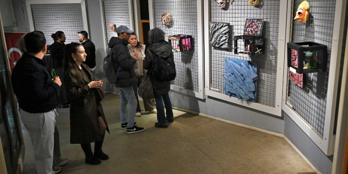 Bir sanat galerisinde veya sergi salonunda, duvarlara asılı metal ızgaralar üzerinde tiyatro temalı maskeler, maketler ve çeşitli sanat eserlerini inceleyen bir grup insan. Katılımcılar ayakta durarak eserlere bakıyor ve kendi aralarında sohbet ediyor. Salonun sol tarafında bir Türk bayrağı yer alıyor.