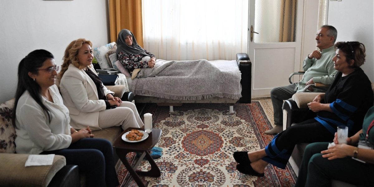 Ev ortamında, yatakta istirahat eden başörtülü yaşlı bir kadını ziyaret eden beş kişilik bir grup. Ziyaretçiler karşılıklı koltuklarda oturarak sohbet ediyor, orta sehpada ise ikramlık kurabiyeler bulunuyor.