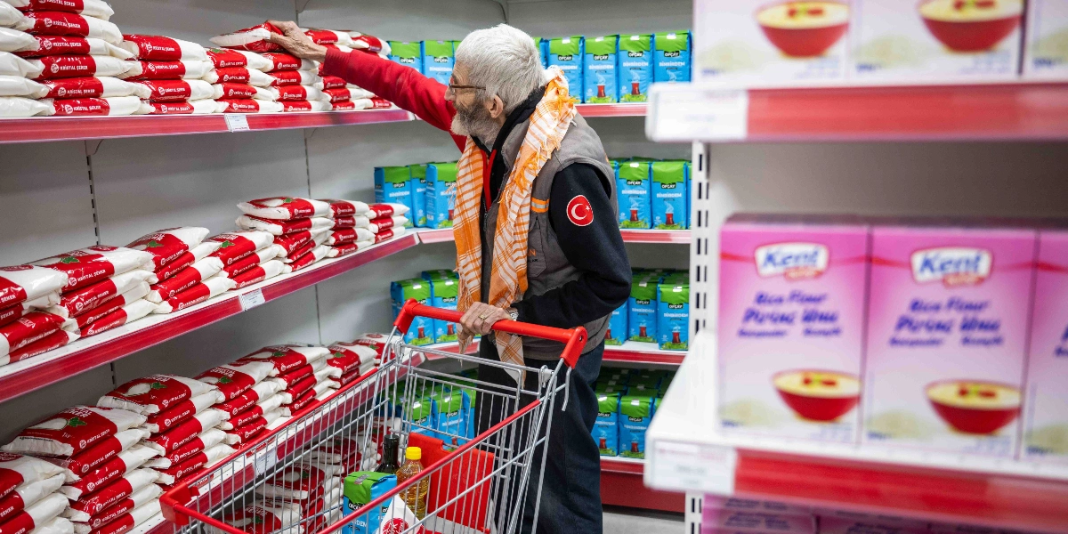 Gri yelekli, beyaz saçlı ve sakallı yaşlı bir adam, market reyonunda alışveriş arabasıyla ilerlerken elini uzatıp raftan bir paket toz şeker alıyor. Adamın yeleğinin kol kısmında Türk bayrağı arması bulunuyor ve boynunda turuncu beyaz çizgili bir şal takılı. Raflar, üzerinde 