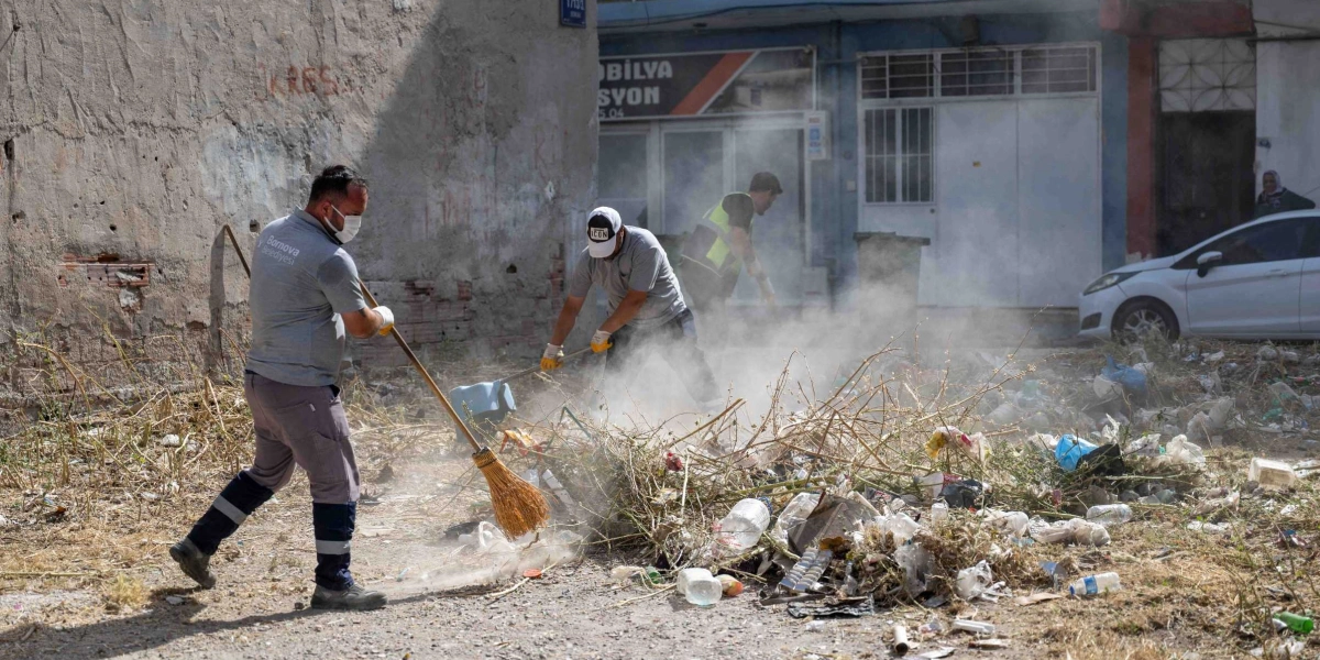 Bornova Belediyesi üniformalı ve maskeli temizlik işçileri, boş bir arazideki çöp ve kuru dal yığınlarını süpürerek temizlik çalışması yapıyor. Çalışma sırasında toz bulutları havaya kalkarken, arka planda dükkanlar ve park halindeki beyaz bir otomobil görülüyor.