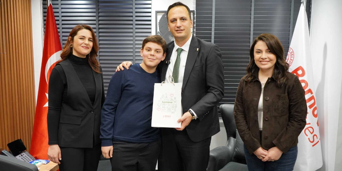 Bornova Belediye Başkanı, yanında duran genç bir öğrenciye üzerinde Bornova Belediyesi logosu bulunan bir hediye çantası veriyor. Yanlarında iki kadın yetkili gülümseyerek eşlik ediyor. Arka planda Türk bayrağı ve Bornova Belediyesi flaması yer alıyor.