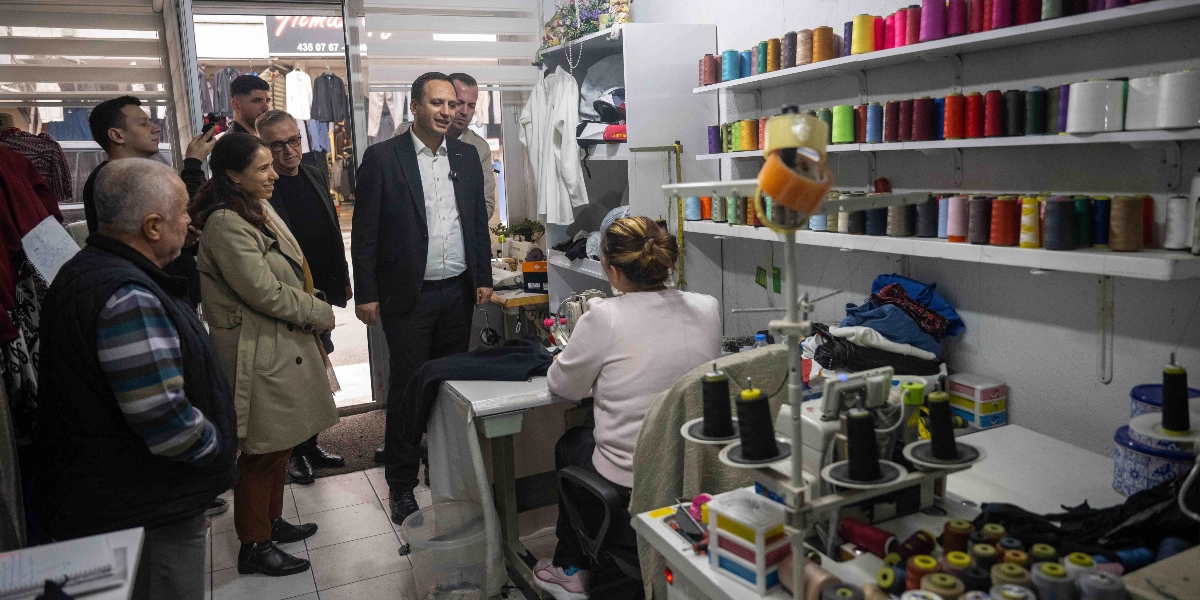 Belediye başkanı ve beraberindeki heyet, Çamdibi'ndeki bir terzi dükkanını ziyaret ederek dikiş makinesinin başında çalışan bir kadınla sohbet ediyor. Dükkanın duvarındaki raflarda çok sayıda renkli dikiş ipliği makarası dizili duruyor.