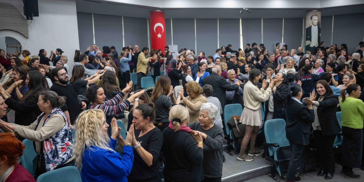 Konferans salonunda bir araya gelmiş kalabalık bir grup insan, karşılıklı durarak birbirlerinin ellerine dokundukları veya alkışladıkları bir etkileşim egzersizi yapıyor. Katılımcıların çoğu ayakta, bazıları ise koltuklarda oturuyor; ortamda neşeli ve katılımcı bir atmosfer hakim. Salonun arka duvarında asılı büyük bir Türk bayrağı ve sağ taraftaki sütunda Mustafa Kemal Atatürk'ün portresi yer alıyor.