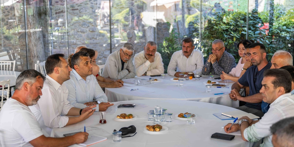 Aydınlık ve cam duvarlı bir mekanda, beyaz örtülü büyük bir yuvarlak masa etrafında toplanmış, aralarında muhtarların da bulunduğu bir grup insan toplantı yapıyor. Masanın üzerinde not kağıtları, kalemler, bardaklar ve atıştırmalık tabakları yer alırken, katılımcılar dikkatle birbirlerini dinliyor ve notlar alıyor.