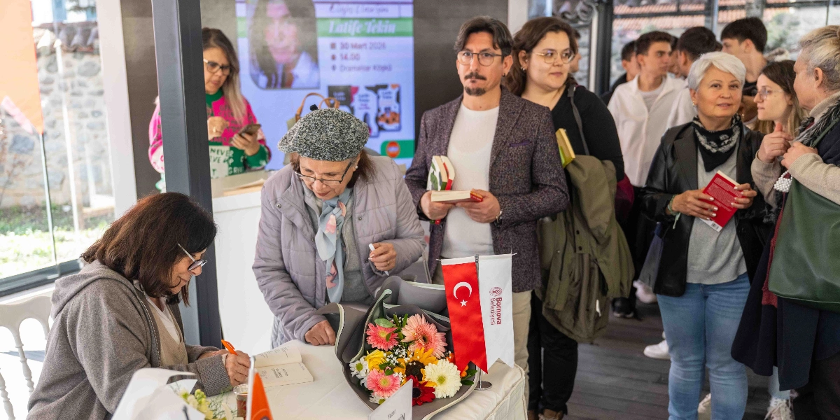 Yazar Latife Tekin, bir masanın başında oturmuş okurları için kitap imzalıyor. Masanın üzerinde bir buket çiçek ve Bornova Belediyesi logolu küçük bir Türk bayrağı bulunuyor. Tekin'in hemen yanında gri şapkalı bir kadın ona bakarken, arkasında sırasını bekleyen çeşitli yaş gruplarından insanlar ellerinde kitaplarla duruyor. Arka planda ise üzerinde Latife Tekin'in fotoğrafı ve etkinlik bilgilerinin yer aldığı bir afiş görünüyor.