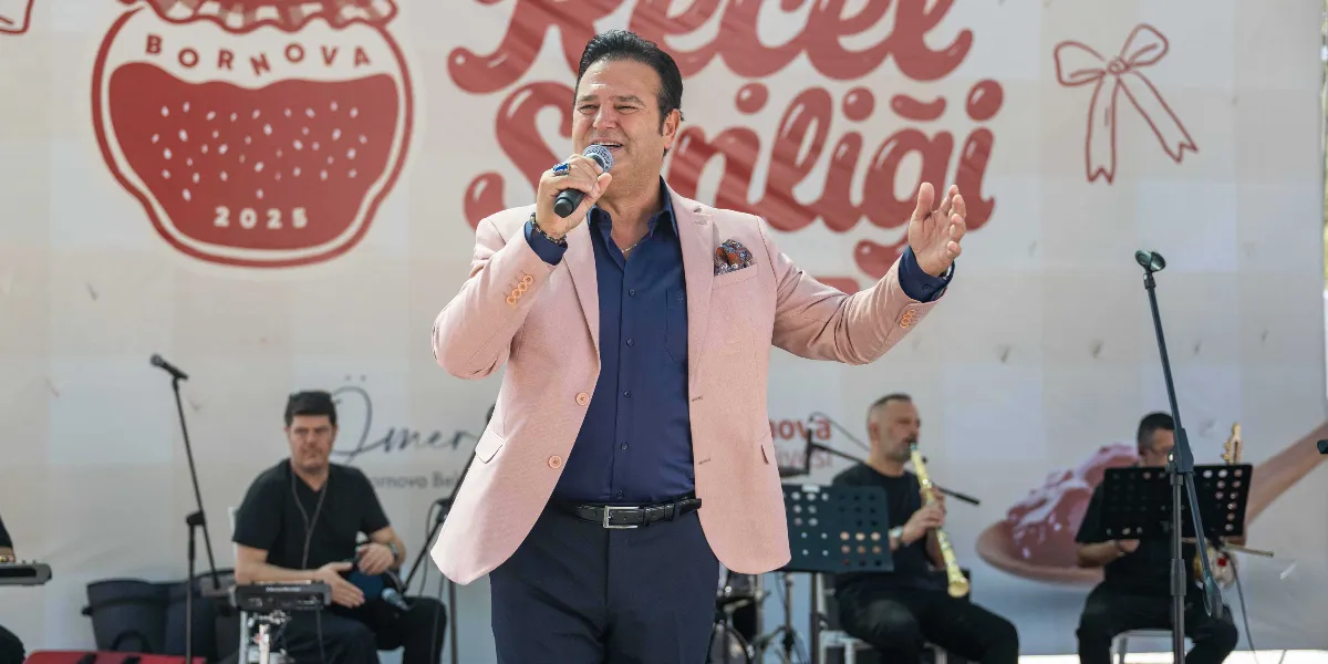 Lacivert gömlek ve üzerine açık pembe bir ceket giymiş bir erkek, elinde mikrofonla sahnede şarkı söylüyor. Arka planda 