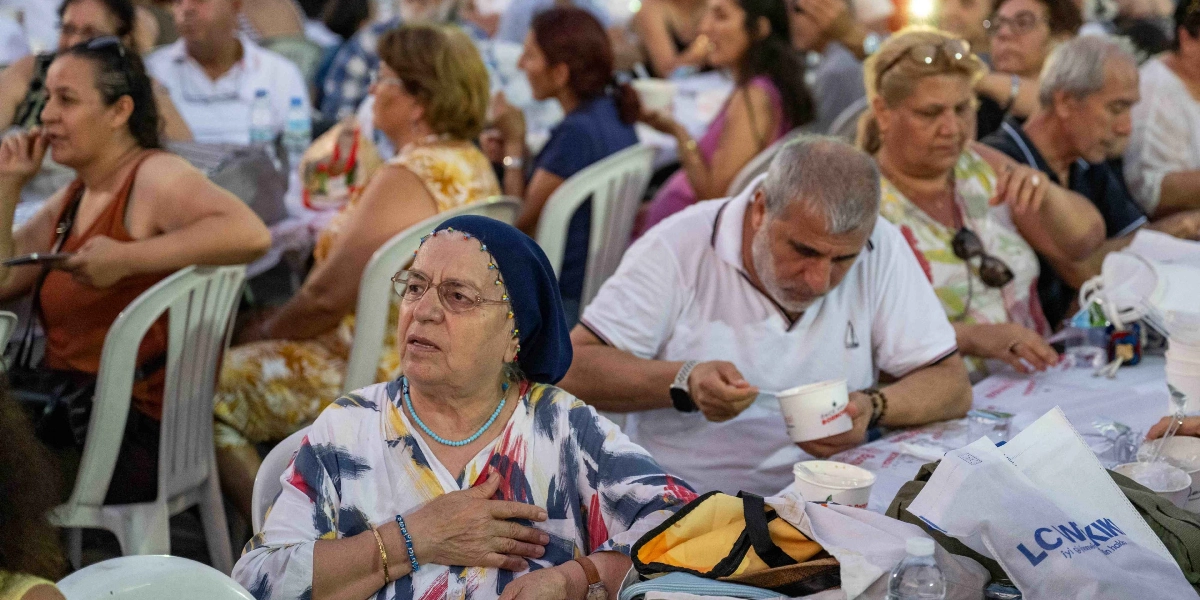 Açık havada uzun masalarda yan yana oturan kalabalık bir grup insan, önlerindeki plastik kaplardan yemek yiyor. Ön planda, başında lacivert bir başörtüsü, üzerinde desenli bir bluz olan ve elini göğsüne koymuş yaşlı bir kadın dikkat çekiyor. Hemen yanında beyaz tişörtlü, orta yaşlı bir adam elinde plastik bir kaseyle yemeğini yiyor. Masaların üzerinde Bornova Belediyesi logolu yemek kapları ve su şişeleri bulunuyor.