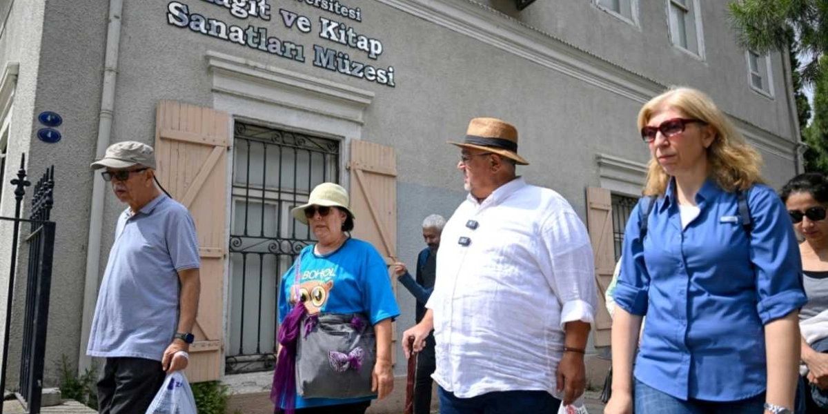 Bornova'daki Kağıt ve Kitap Sanatları Müzesi binasının önünde yürüyen bir grup insan. Grupta güneş gözlüklü ve şapkalı kişiler ile beyaz gömlekli, fötr şapkalı bir erkek ve mavi gömlekli bir kadın ön planda yer alıyor. Müze binasının gri taş duvarında 