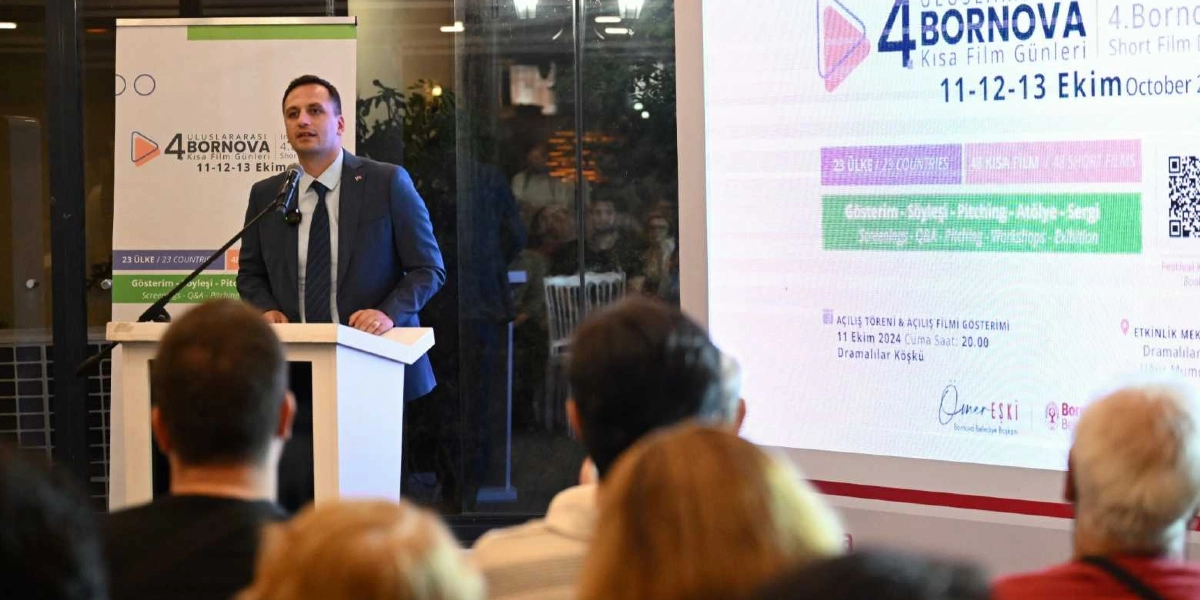 Koyu renk takım elbiseli bir konuşmacı, kürsüde mikrofon başında izleyicilere hitap ediyor. Konuşmacının arkasında 4. Uluslararası Bornova Kısa Film Günleri'ne ait detayların yer aldığı büyük bir ekran ve bilgilendirme panosu bulunuyor. Ekranda 11-12-13 Ekim tarihlerinde gerçekleşecek etkinlikte 23 ülkeden 48 kısa filmin gösterileceği, ayrıca söyleşi, pitching, atölye ve sergi gibi faaliyetlerin yer alacağı bilgisi ile Dramalılar Köşkü'ndeki açılış törenine dair detaylar okunabiliyor. Ön planda ise konuşmayı dinleyen izleyicilerin başları görünüyor.