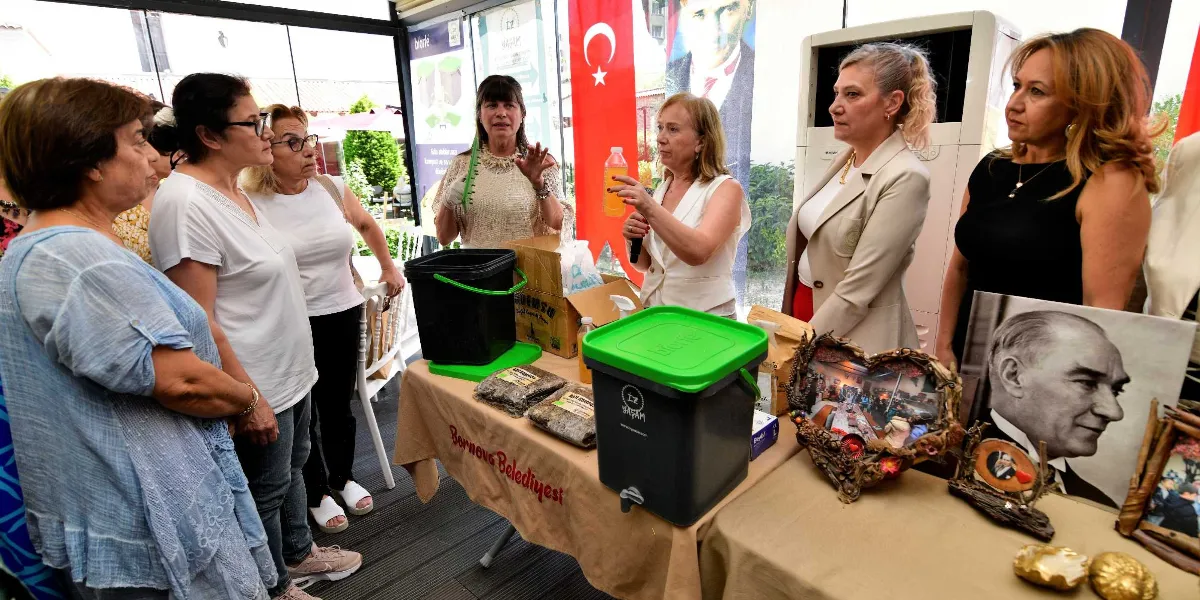 Bornova Belediyesi tarafından düzenlenen bir etkinlikte, bir grup kadın katılımcı, kompost yapımı hakkında bilgi veren bir sunumu dinliyor. Masanın üzerinde yeşil kapaklı kompost kovaları, organik atıklar ve üzerinde Mustafa Kemal Atatürk'ün fotoğrafının bulunduğu, kalp şeklinde süslenmiş bir çerçeve yer alıyor. Arka planda Türk bayrağı ve Atatürk posteri görülüyor.
