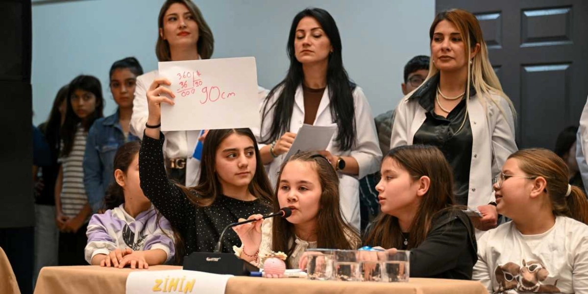 Bir etkinlikte, masada oturan ve mikrofonla konuşan bir kız çocuğu, üzerinde matematiksel bir bölme işlemi ve 