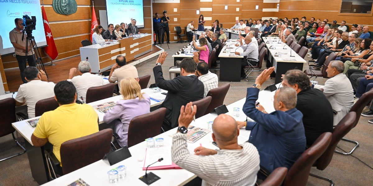 Bornova Belediyesi'nin Eylül ayı olağan meclis toplantısında, katılımcıların büyük çoğunluğunun oylama yapmak için ellerini kaldırdığı geniş katılımlı bir oturum. Salonun ön kısmında başkanlık divanı, arkasında ise sıralar halinde oturan meclis üyeleri ve izleyiciler yer alıyor. Sol tarafta bir kameraman çekim yaparken, arka plandaki ekranda toplantı başlığı yansıtılıyor.
