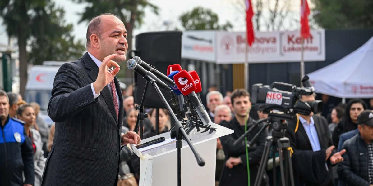 Bornova Belediye Başkanı, önünde mikrofonların bulunduğu bir kürsüde konuşma yapıyor. Arka planda Bornova Belediyesi'ne ait bir yapı ve etkinlik alanında bulunan kalabalık bir grup yer alıyor. Sağ tarafta ise çekim yapan bir kameraman ve basın mensupları görülüyor.