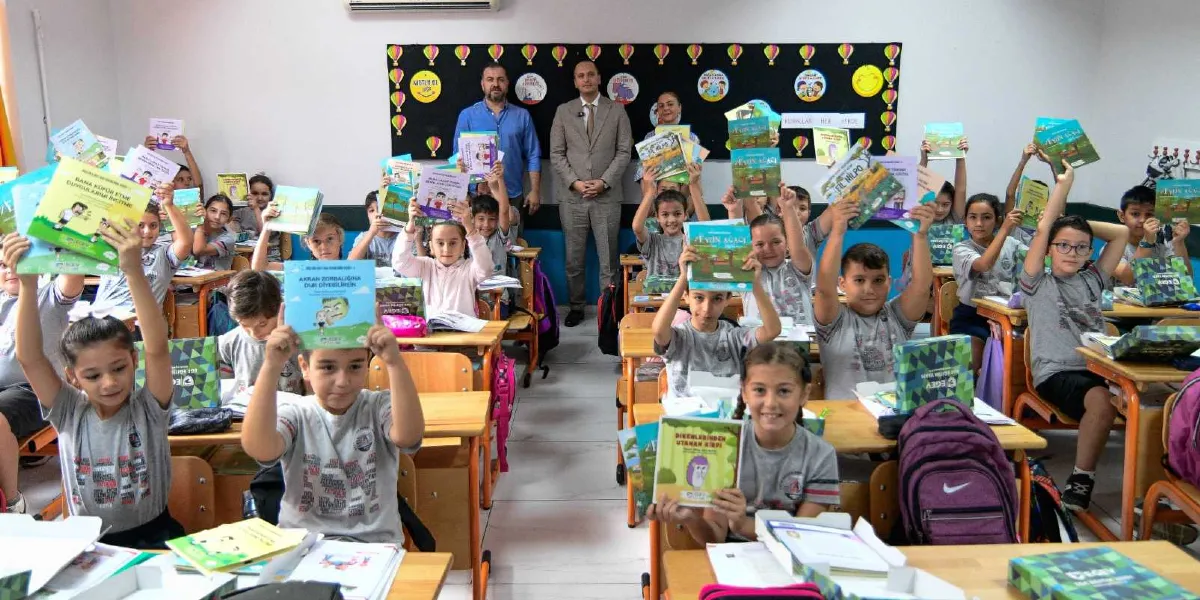 Sınıf ortamında sıralarında oturan ilkokul çağındaki çocuklar, Bornova Belediyesi tarafından hediye edilen kitapları havaya kaldırarak neşeyle poz veriyor. Çocukların önündeki sıralarda kırtasiye malzemeleri ve kitaplar bulunuyor. Sınıfın arka tarafında ise üç yetişkin, öğrencilerin arkasında ayakta durarak onlara eşlik ediyor. Arka duvarda üzerinde 