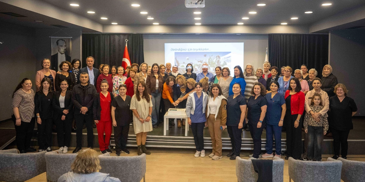 Modern bir konferans salonunda, sahne üzerinde bir araya gelmiş kalabalık bir grup insan, bir kutlama veya etkinlik kapsamında pasta kesiyor. Grubun merkezinde yer alan birkaç kişi, küçük bir masanın üzerindeki pastayı birlikte keserken, diğerleri onları izleyerek gülümsüyor. Arka planda Türk bayrağı, Atatürk portresi ve üzerinde 