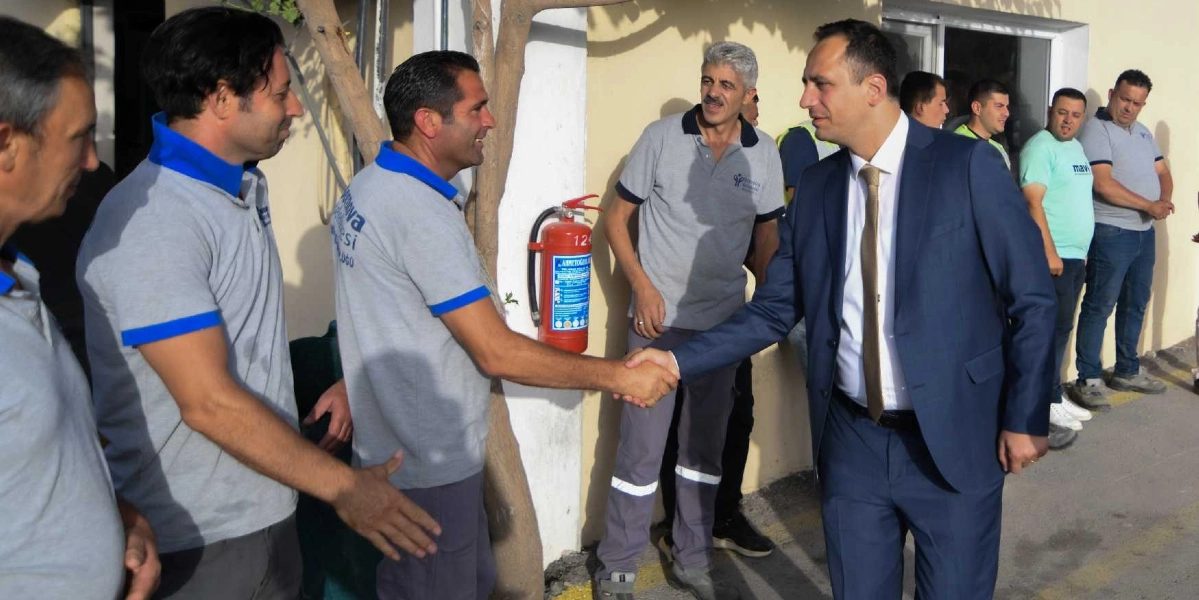 Koyu mavi takım elbiseli ve kravatlı bir yetkili, üzerinde Bornova Belediyesi yazılı gri üniformalar giyen çalışanlarla tokalaşıyor. Dış mekanda, bir binanın önünde gerçekleşen bu buluşmada, çalışanlar ve yetkili gülümseyerek birbirini selamlıyor. Arka planda duvara asılı bir yangın tüpü ve diğer çalışanlar yer alıyor.