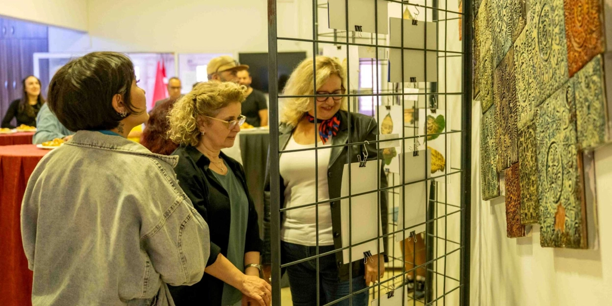 İç mekan bir sanat sergisinde, metal bir ızgara panoya mandallarla tutturulmuş kağıtları ve yanındaki kabartmalı desenlere sahip seramik karoları inceleyen üç kadın. Ön planda sırtı dönük, kot ceketli genç bir kadın, yanında gözlüklü ve kıvırcık saçlı orta yaşlı bir kadın ile beyaz tişört ve siyah deri ceket giymiş sarışın bir kadın sergilenen eserlere dikkatle bakıyor. Arka planda ise bulanık şekilde diğer katılımcılar ve kırmızı örtülü masalar görülüyor.