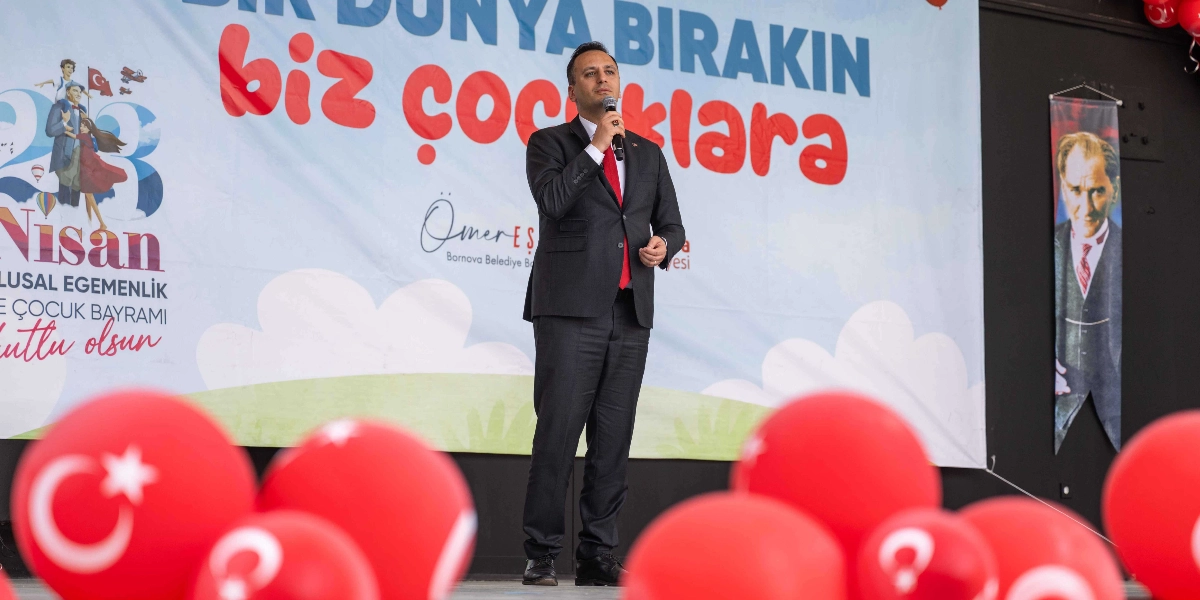 Koyu renk takım elbiseli ve kırmızı kravatlı bir erkek, 23 Nisan Ulusal Egemenlik ve Çocuk Bayramı temalı bir sahne dekorunun önünde elinde mikrofonla konuşma yapıyor. Arka planda 