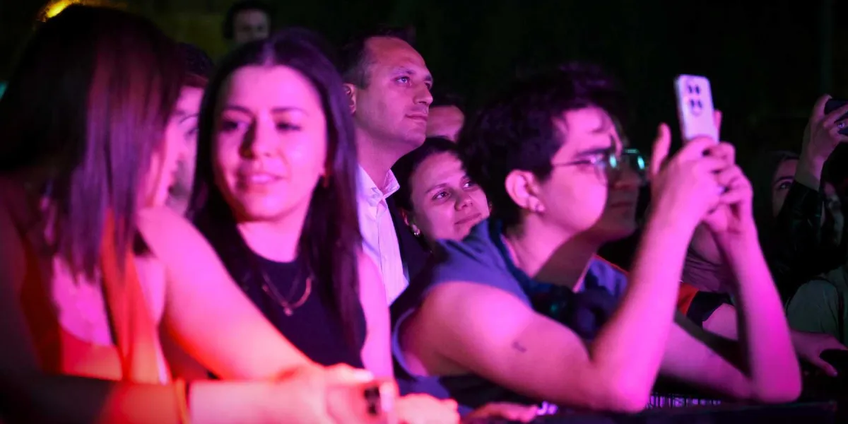 Gece vakti düzenlenen bir etkinlikte, sahneye odaklanmış kalabalık bir grup genç. Ön planda, mor ve pembe sahne ışıklarıyla aydınlanan, gülümseyen genç bir kadın ve yanında telefonuyla çekim yapan gözlüklü bir genç yer alıyor. Arka planda ise farklı yaşlardan insanlar dikkatle sahneyi izliyor.