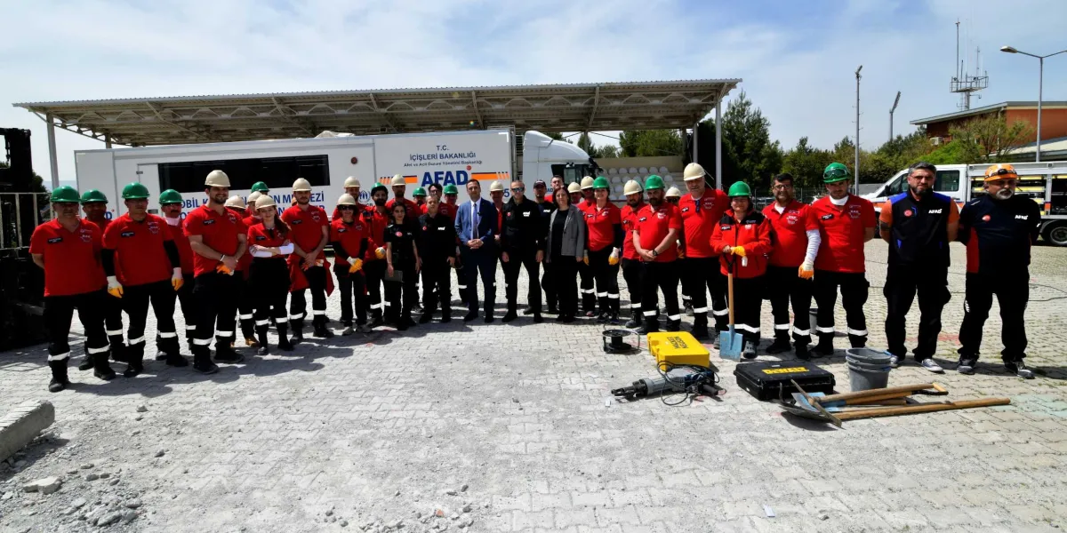 Açık havada, arkalarında üzerinde AFAD logolu bir tırın bulunduğu alanda yan yana dizilmiş, kırmızı tişörtlü, baretli ve iş kıyafetli Bornova Belediyesi arama kurtarma ekibi üyeleri. Ekibin önünde kurtarma çalışmalarında kullanılan kürek, kırıcı delici alet ve taşıma çantaları gibi ekipmanlar yer alıyor. Grubun ortasında takım elbiseli bir yetkili ve yanında siyah ceketli bir kadın bulunuyor.