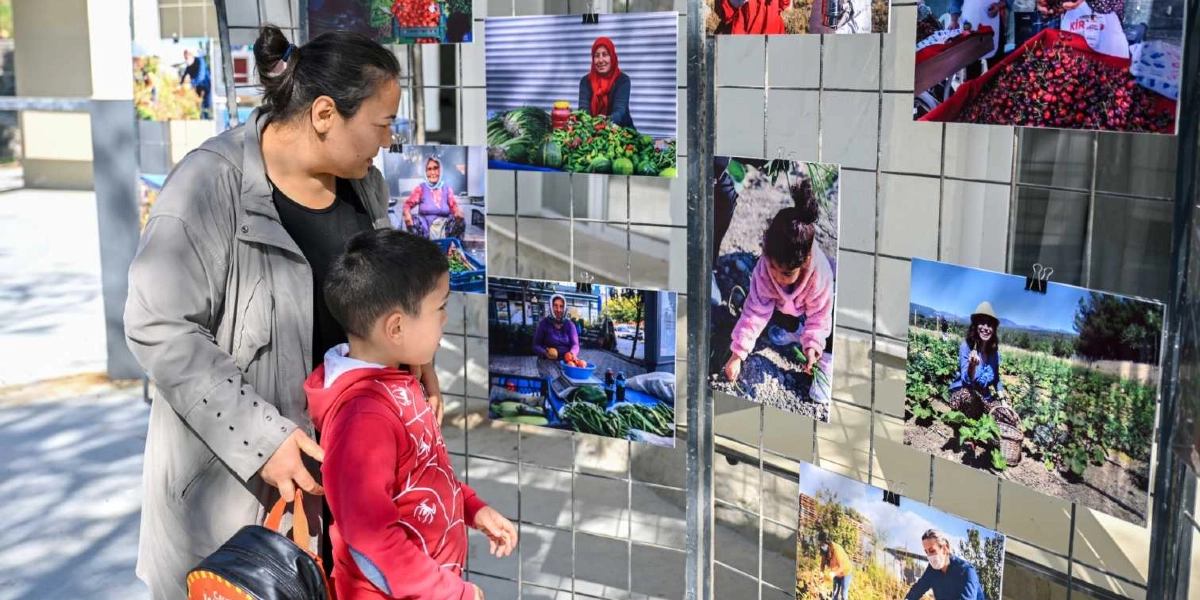 Gri ceketli bir kadın ve yanındaki kırmızı sweatshirtlü küçük bir çocuk, metal bir tel ızgara üzerine asılmış fotoğraflardan oluşan bir sergiyi inceliyor. Fotoğraflarda tarlada çalışan, ürünlerini pazarda satan kadınlar ve bahçeyle ilgilenen bir çocuk yer alıyor.