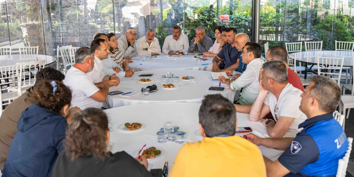 Aydınlık ve cam duvarlı bir mekanda, beyaz örtülü büyük yuvarlak masaların etrafında toplanmış, aralarında muhtarların ve yerel yetkililerin bulunduğu kalabalık bir grup toplantı yapıyor. Katılımcılar masalara dağılmış şekilde oturuyor, bazıları not alıyor, bazıları ise birbirini dinleyerek fikir alışverişinde bulunuyor. Masaların üzerinde su bardakları, küçük atıştırmalık tabakları, kalemler ve not defterleri yer alıyor. Ön planda sarı tişörtlü bir kişi ve zabıta üniformalı bir görevli sırtları dönük şekilde otururken, masanın diğer tarafında farklı yaş ve cinsiyetten insanlar bir arada görünüyor.