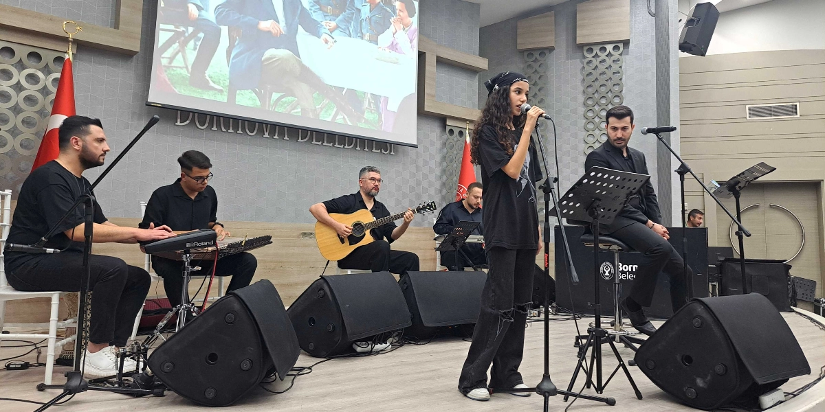 Bornova Belediyesi salonunda düzenlenen bir etkinlikte, sahnede canlı müzik performansı sergileyen beş kişilik bir grup yer alıyor. En önde mikrofonla şarkı söyleyen genç bir kız, arkasında ise kanun, akustik gitar ve klavye çalan müzisyenler ile bir başka vokalist bulunuyor. Sahnenin arka planındaki dev ekranda nostaljik bir fotoğraf yansıtılırken, yanlarda Türk bayrakları ve Bornova Belediyesi'nin logosu görülüyor.