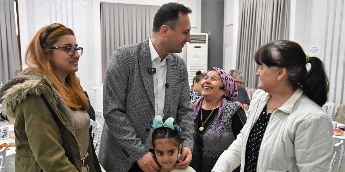 Gri ceketli bir erkek, bir dayanışma yemeğinde ailelerle bir araya gelerek sohbet ediyor. Yanında gözlüklü genç bir kadın, başında pembe desenli bir eşarp olan gülen yaşlı bir kadın ve saçında parlak mavi bir fiyonk bulunan küçük bir kız çocuğu yer alıyor. Grubun karşısında ise beyaz ceketli bir kadın onlarla etkileşimde bulunuyor. Arka planda yemek masaları ve diğer katılımcılar görülüyor.