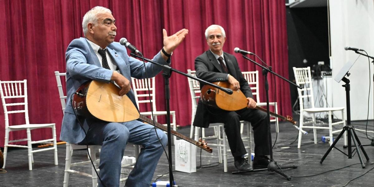 Sahne üzerinde, arkalarında bordo renkli bir perde bulunan iki müzisyen beyaz sandalyelerde oturarak bağlama çalıyor. Öndeki müzisyen açık mavi bir takım elbise giymiş, elini havaya kaldırarak konuşuyor ve bağlamasını çalıyor. Yanında, koyu renk takım elbiseli diğer müzisyen ise elinde bağlamasıyla ona eşlik ediyor. Her iki müzisyenin önünde mikrofonlar ve ayaklıklar bulunuyor.