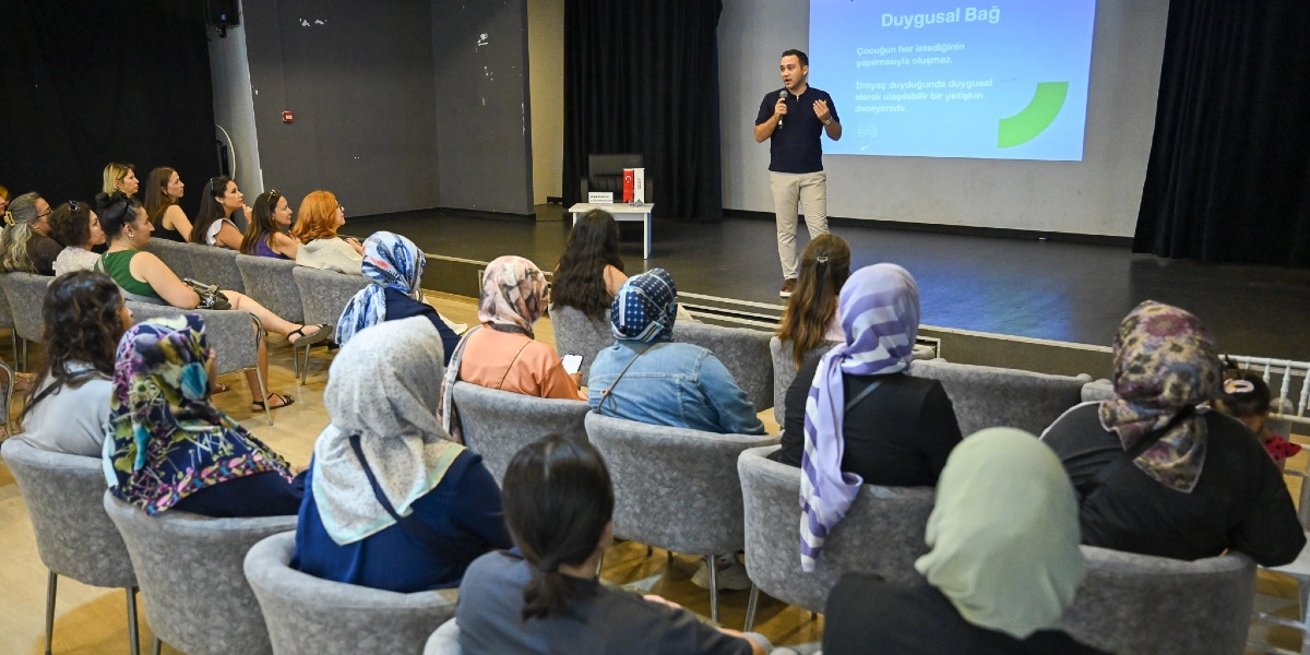 Konferans salonunda sahne üzerinde elinde mikrofonla konuşma yapan bir uzman, çocuk eğitimi üzerine sunum yapıyor. Arkasındaki yansıtıcı ekranda 