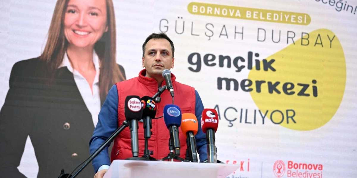 Kırmızı yelek giymiş bir erkek, önünde çok sayıda mikrofonun bulunduğu bir kürsüde konuşma yapıyor. Arka planda Bornova Belediyesi Gülşah Durbay Gençlik Merkezi açılışını duyuran bir afiş ve üzerinde Gülşah Durbay'ın fotoğrafı yer alıyor.