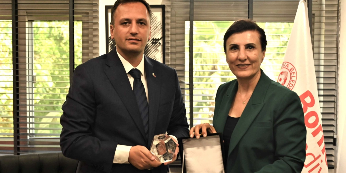 Koyu renk takım elbiseli bir erkek ve yeşil ceketli bir kadın, ellerinde kristal bir ödül plaketiyle yan yana duruyor. Arka planda Bornova Belediyesi bayrağı ve jaluzi perdeli pencereler yer alıyor.