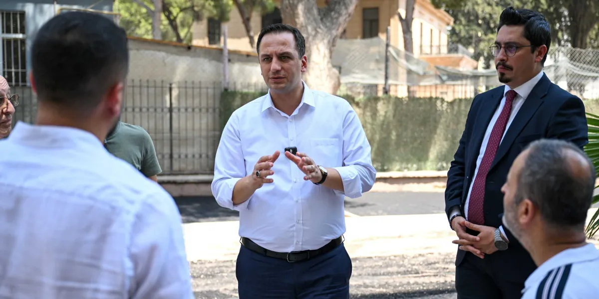 Beyaz gömlekli bir yetkili, dış mekanda bir grup insanla konuşurken elleriyle açıklama yapıyor. Yanında lacivert takım elbiseli ve kırmızı kravatlı bir kişi duruyor. Arka planda ise ağaçlar, demir parmaklıklı bir duvar ve tarihi bir yapı binası görülüyor.