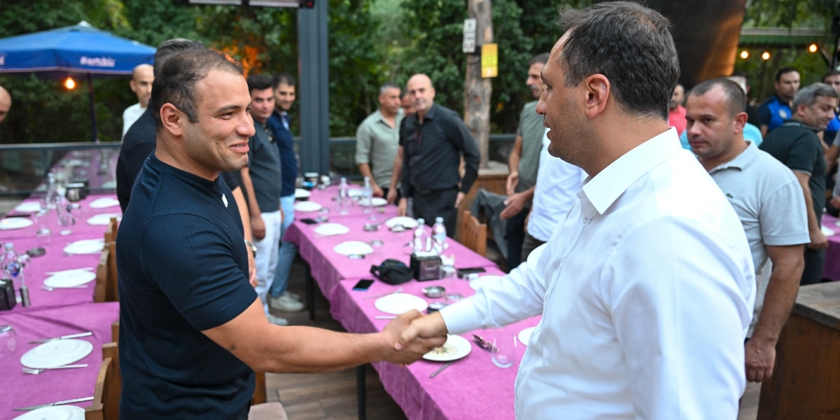 Bornova Belediye Başkanı Ömer Eşki, açık havada düzenlenen bir yemek organizasyonunda zabıta personeliyle tokalaşarak selamlaşıyor. Arka planda mor örtülü uzun yemek masaları ve çevrede ayakta duran diğer belediye çalışanları yer alıyor.