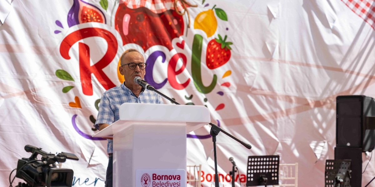 Gözlüklü ve kareli kısa kollu gömlek giyen bir erkek, üzerinde Bornova Belediyesi logosu bulunan beyaz bir kürsüde mikrofon başında konuşma yapıyor. Arka planda, üzerinde büyük harflerle 