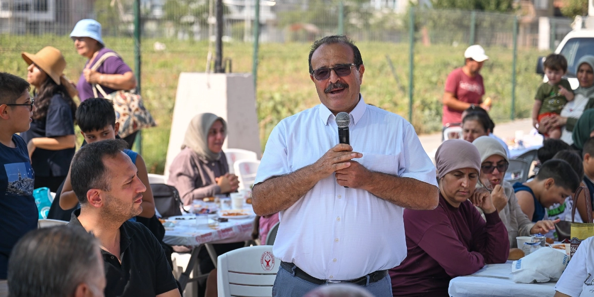 Açık havada düzenlenen bir etkinlikte, beyaz kısa kollu gömlek giymiş, gözlüklü ve bıyıklı bir adam elinde mikrofonla konuşma yapıyor. Adamın arkasında ve çevresinde, masalarda oturan, yemek yiyen ve birbirleriyle sohbet eden çeşitli yaş gruplarından insanlar bulunuyor. Arka planda ise tel örgülerle çevrili geniş bir tarım arazisi ve yerleşim yeri görülüyor.